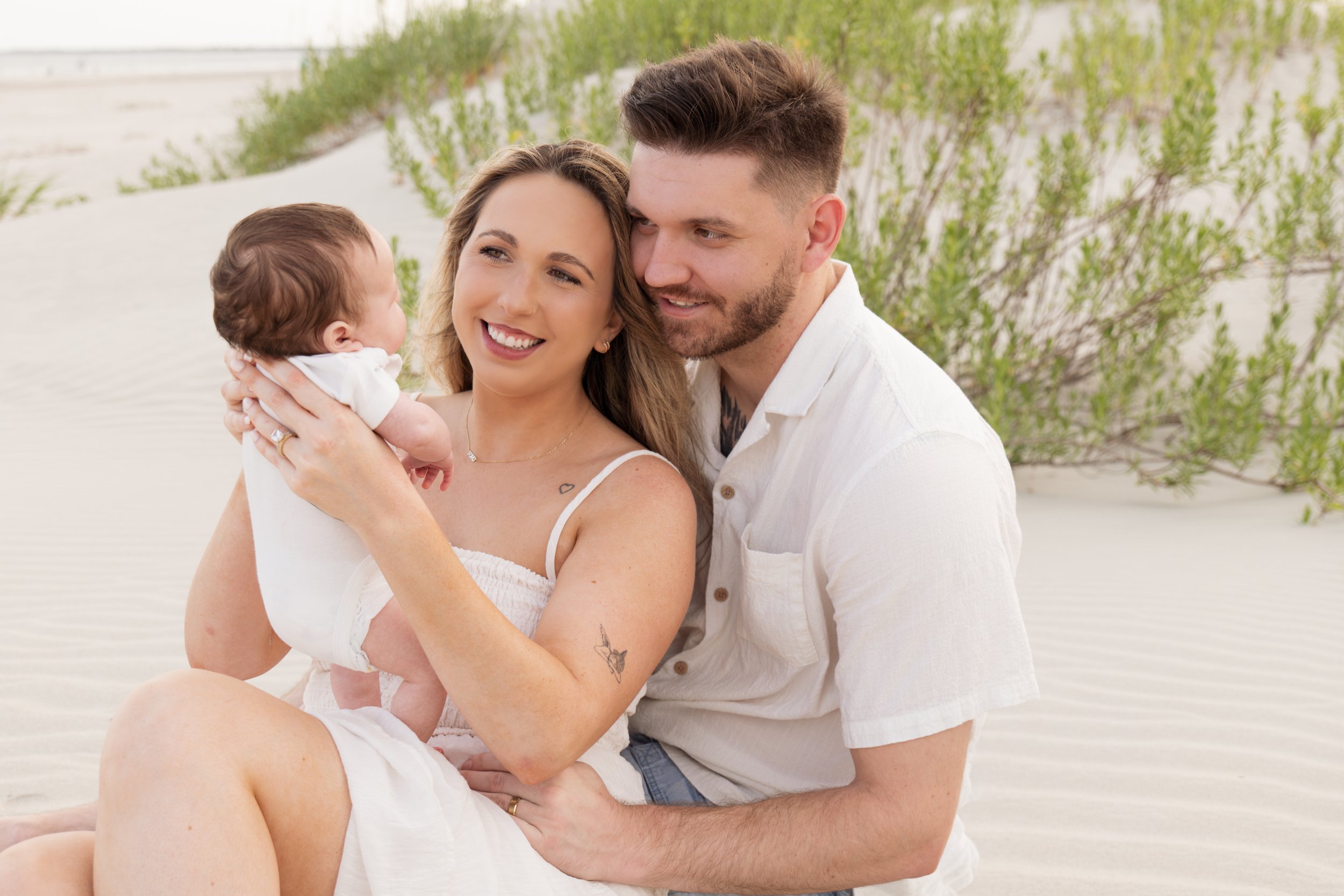 A happy family of three, a woman, a man, and a baby, enjoying quality time together outdoors on a sunny day near white dunes and green bushes.