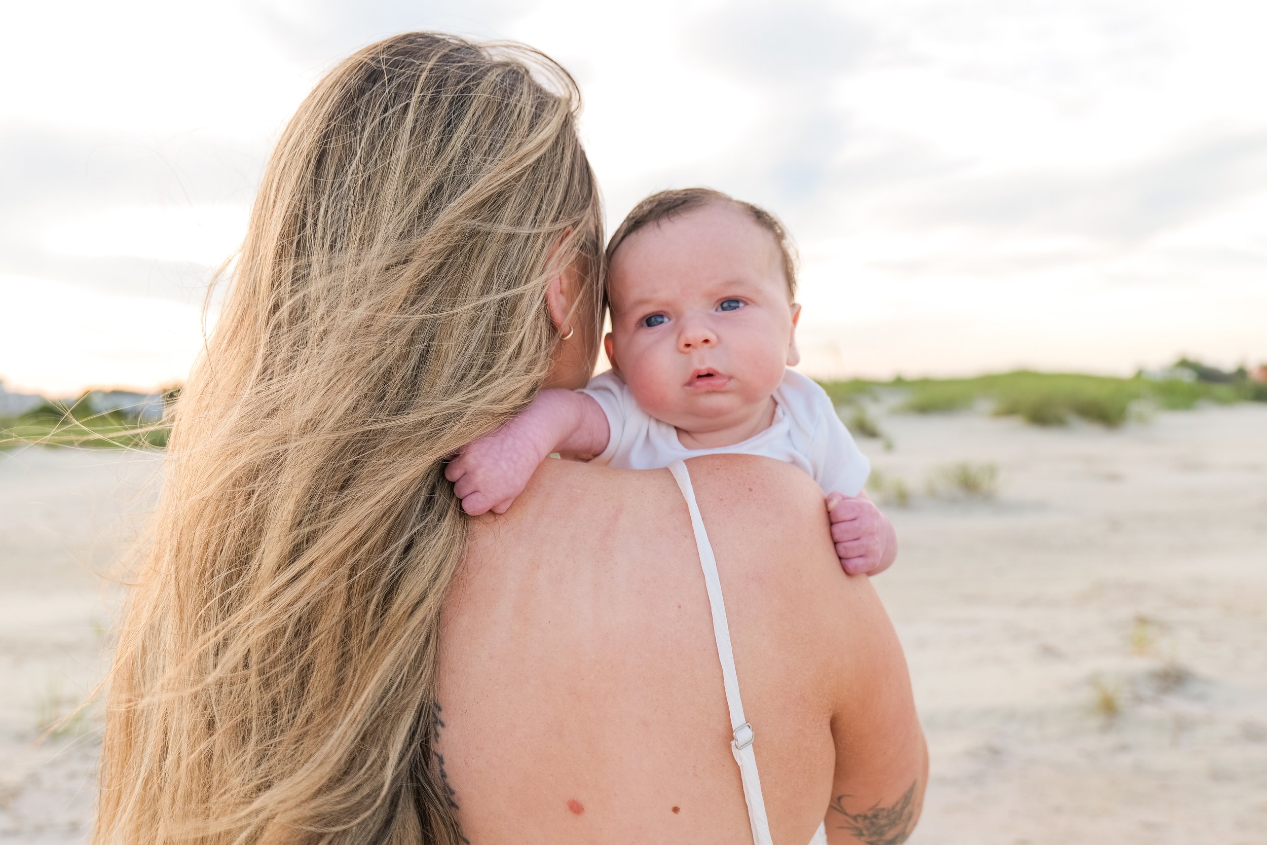 A woman with long blonde hair holding a baby with blue eyes, on a beach with sand and shrubs in the background.
