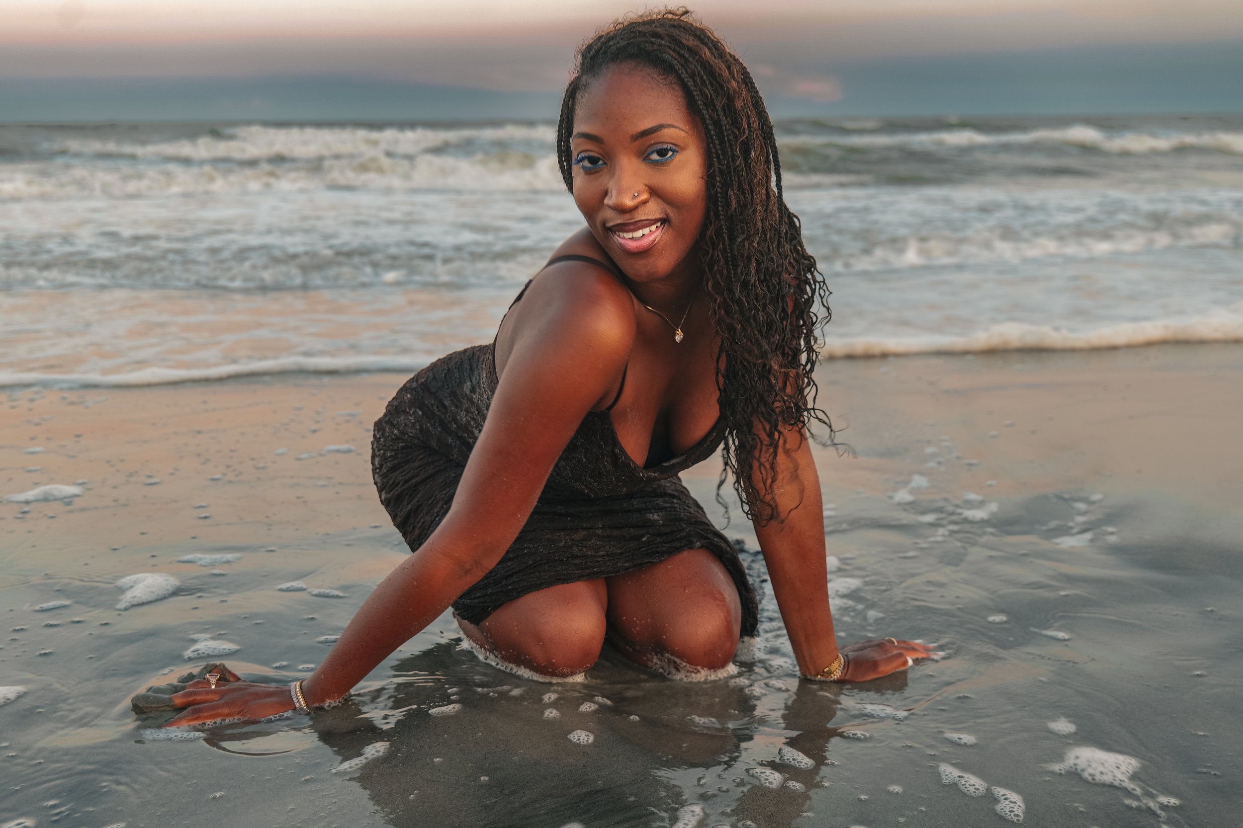 A woman with long curly hair and blue eyeshadow kneels on a sandy beach with waves in the background during sunset, smiling at the camera.