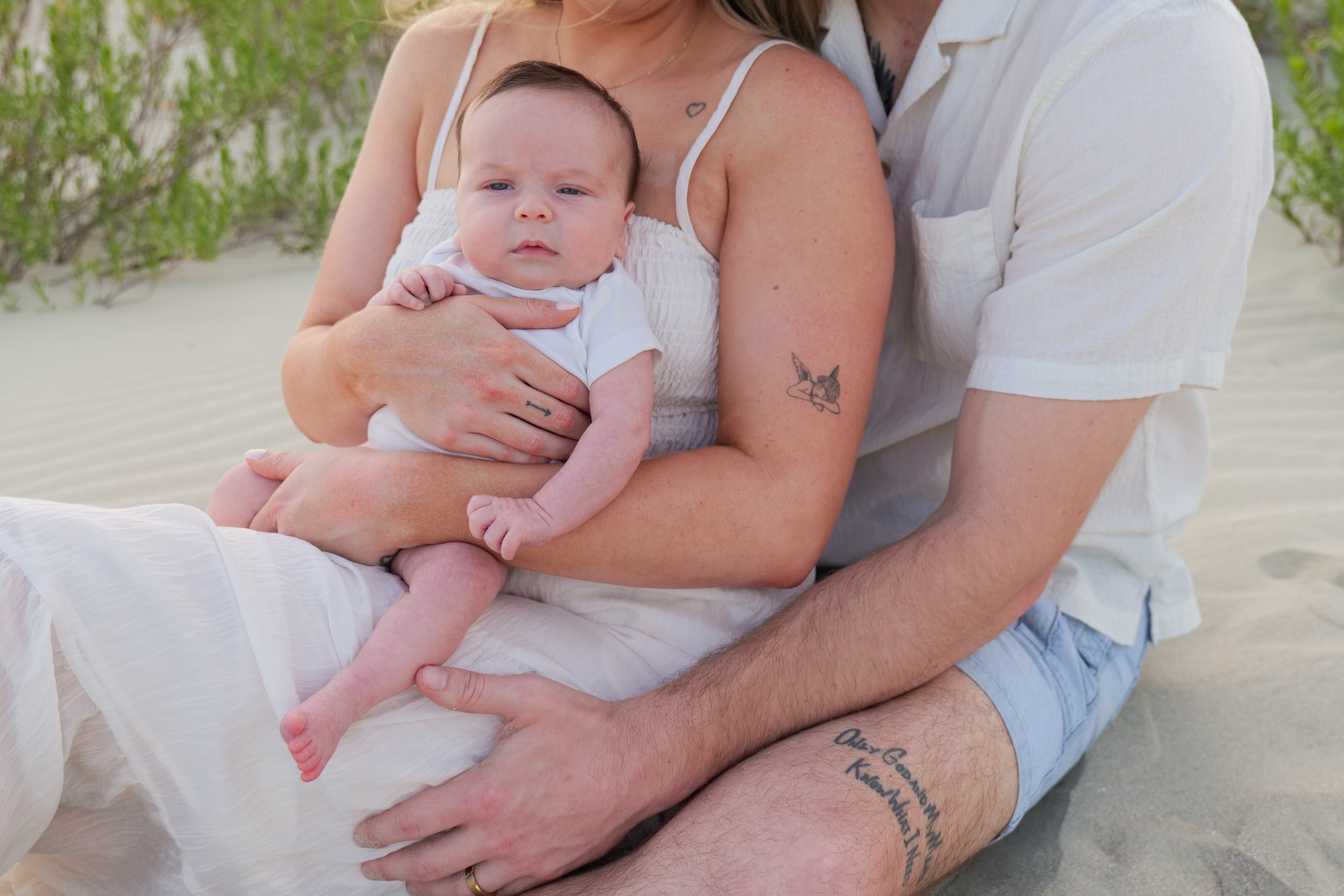 A couple holding a baby outdoors, with the woman wearing a white dress and the man in a white shirt and denim shorts.
