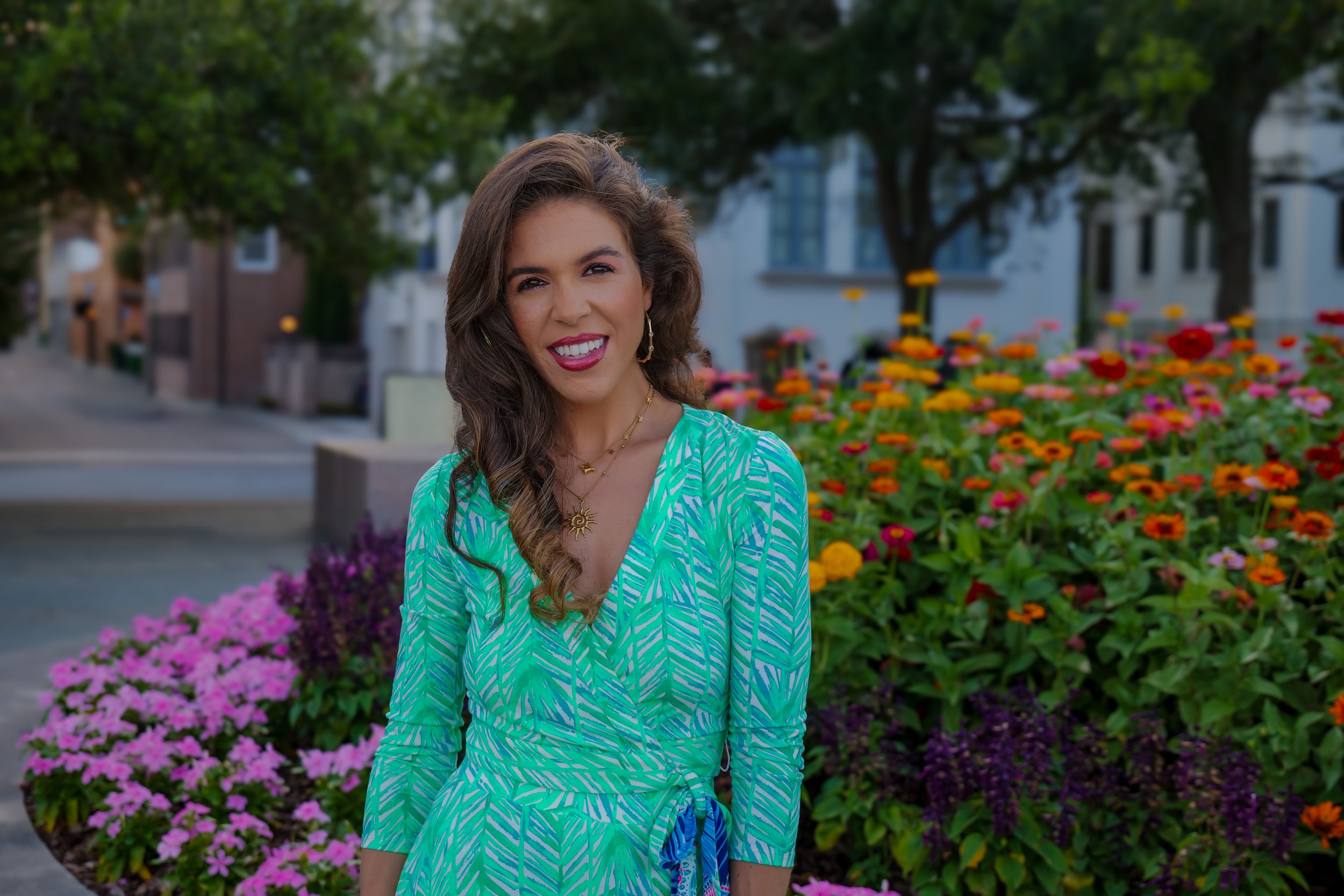 A young woman with long, wavy brown hair wearing a green and blue patterned dress, smiling outdoors with a background of colorful flowers, trees, and residential buildings.
