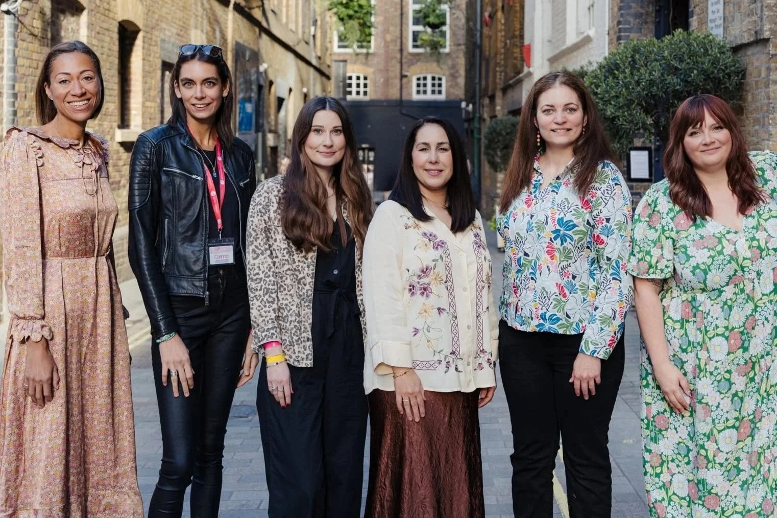 Six women standing outdoors on a city street, smiling at the camera, wearing casual and colorful clothing.