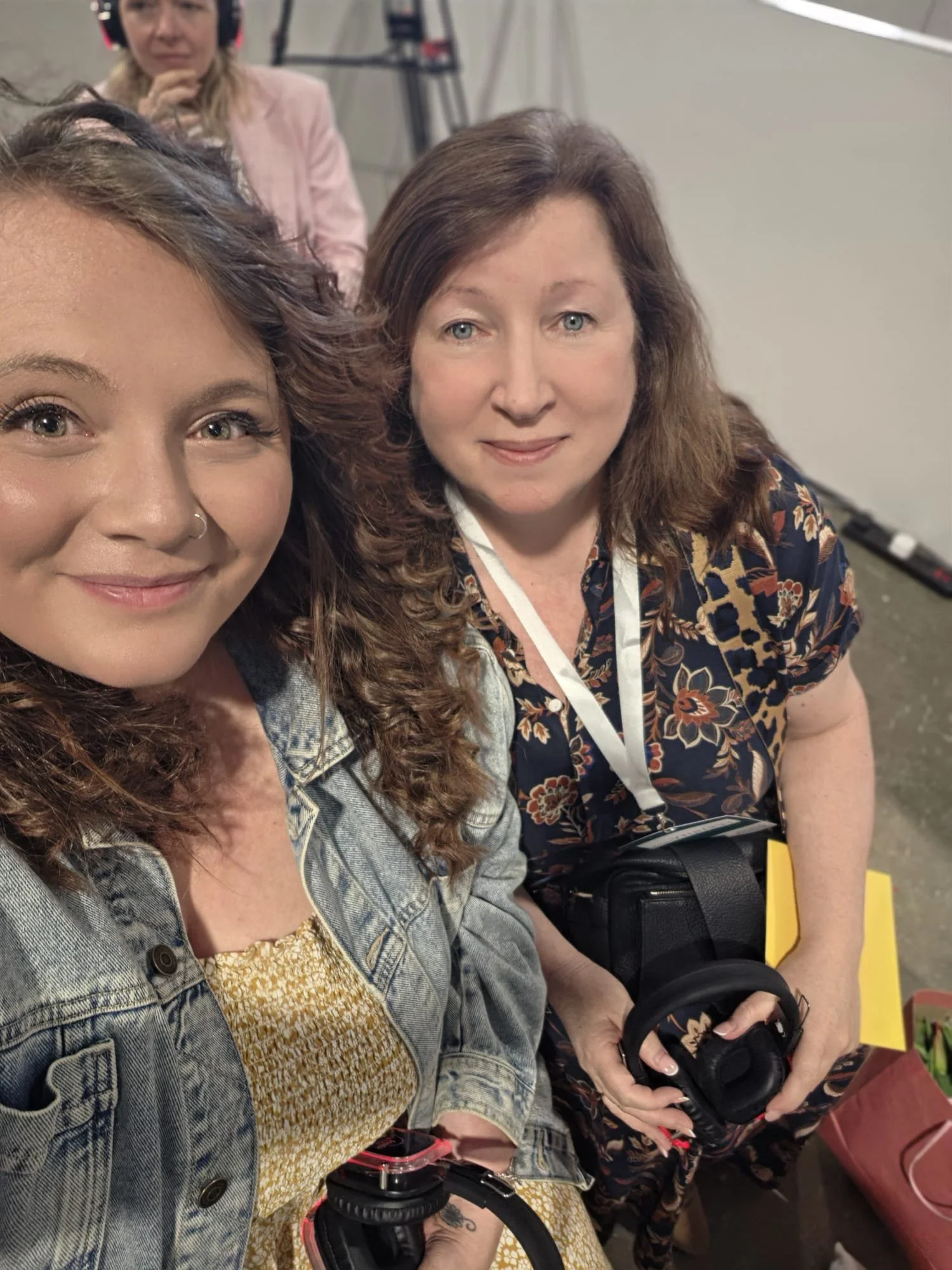 Two women sitting together, one with long, curly brown hair and blue eyes wearing a floral dress, the other with curly hair wearing a denim jacket and yellow patterned top. They are smiling at the camera, with headphones and other people blurred in the background.