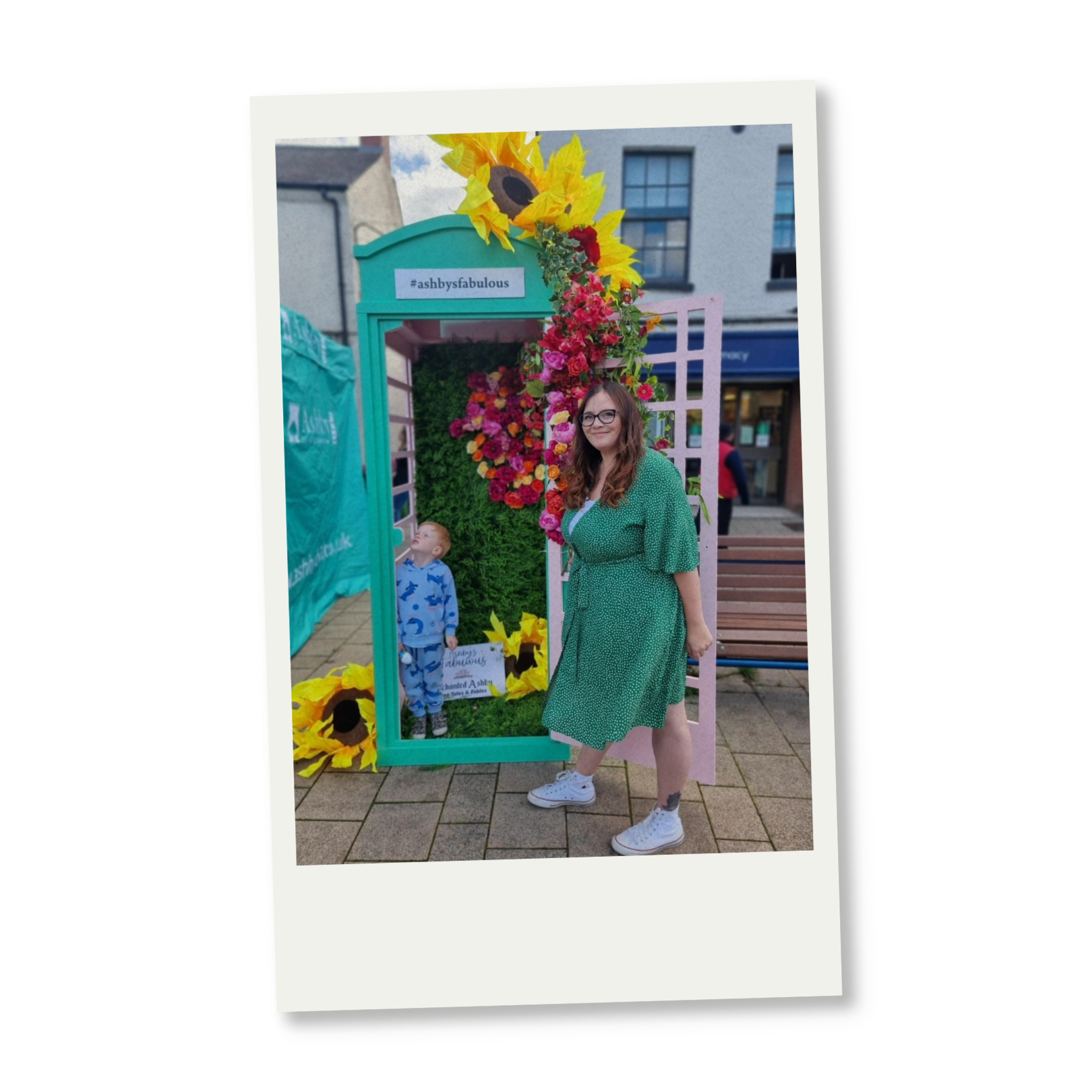 A woman with long brown hair, glasses, wearing a green dress and white sneakers, poses in front of a colorful floral display with a green frame and pink gate. A young boy in pajamas stands nearby, looking up at the flowers. The background shows an outdoor urban setting with buildings and a bench.