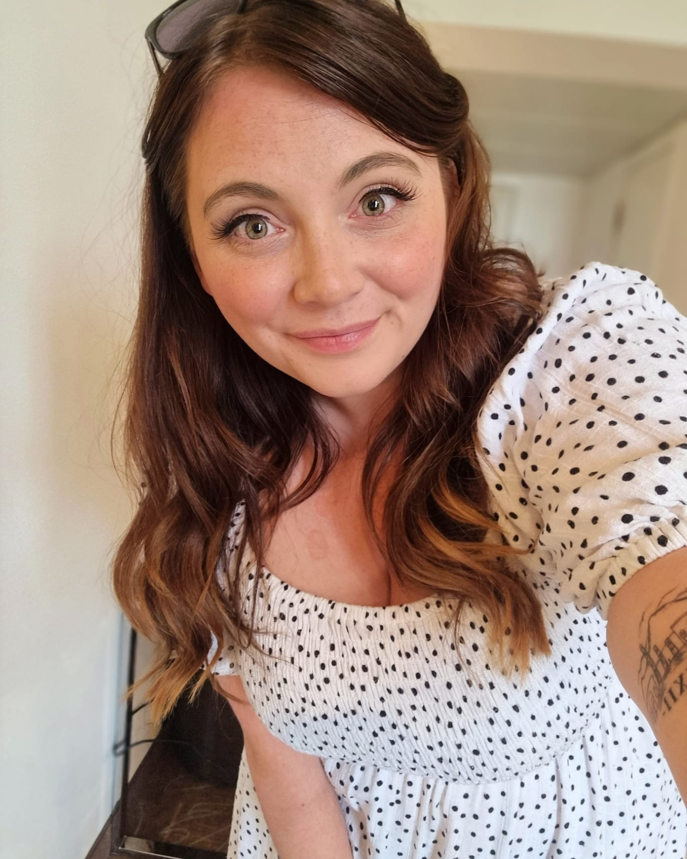 A woman with long, wavy brown hair and green eyes wearing a white dress with small black polka dots, smiling and looking at the camera.