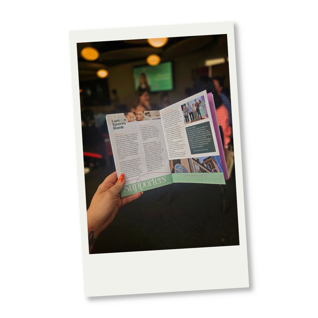A person holding a brochure at an indoor event, with a blurred background of people and a screen.