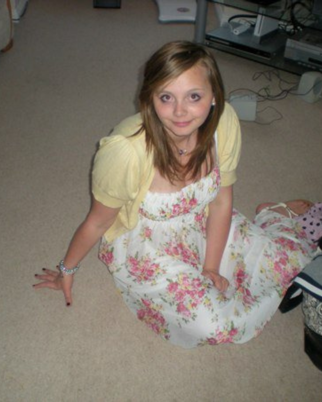 A young woman with light brown hair sitting on a beige carpeted floor, wearing a floral dress with pink and yellow flowers and a yellow jacket. She is looking up at the camera with a slight smile, and has her left hand resting on the floor.