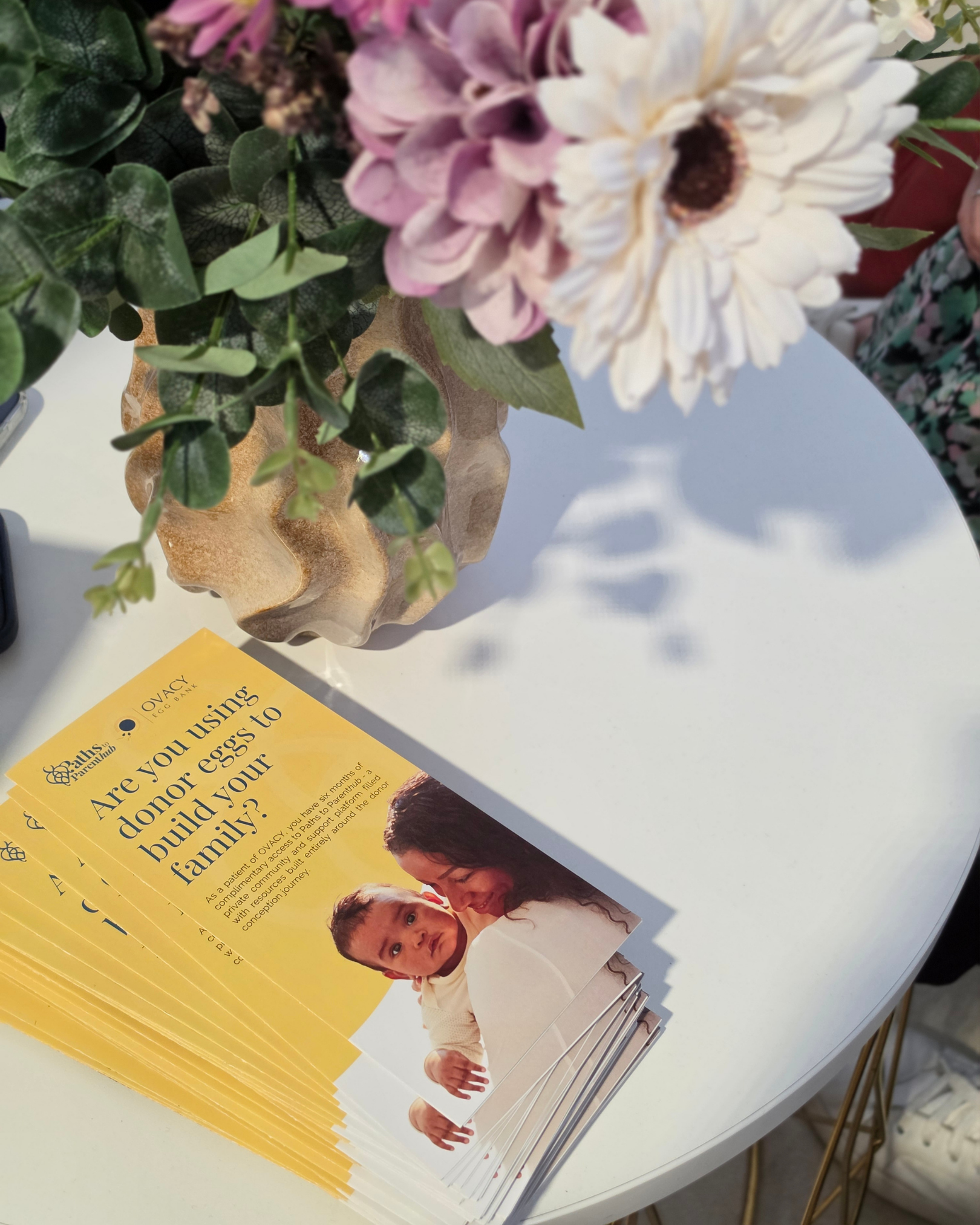 A stack of yellow pamphlets on a white table with a close-up of a family on the front cover, and part of a plant with green leaves and pink and white flowers in the background.