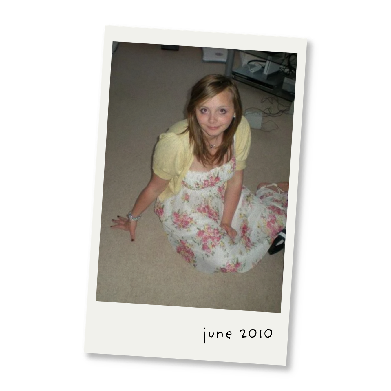 A young girl with brown hair sitting on the floor wearing a floral dress and a yellow cardigan, with a computer and wires in the background, and the text 'June 2010' at the bottom.