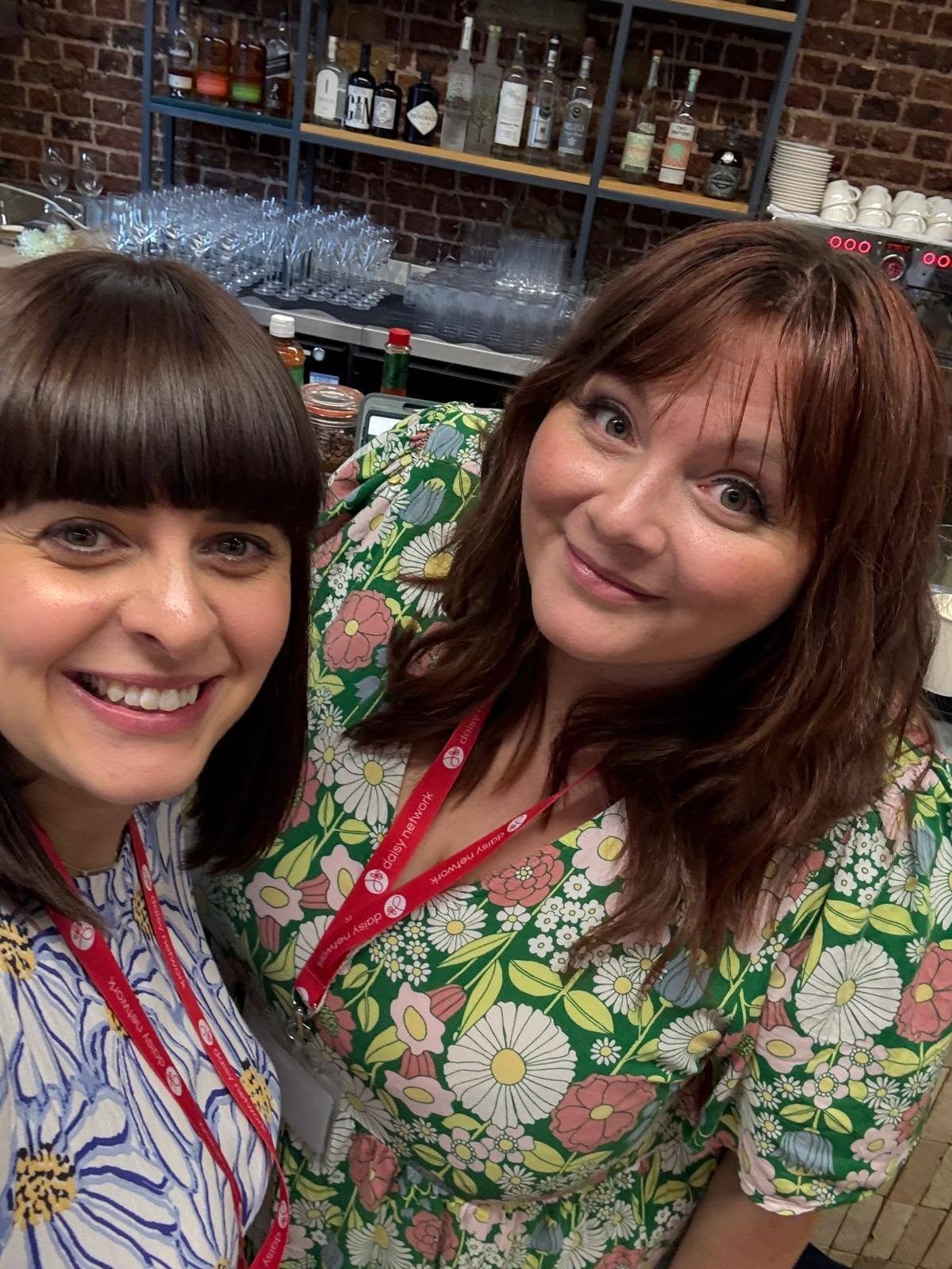 Two women smiling for a selfie in a bar or restaurant, with shelves of bottles and glasses in the background.