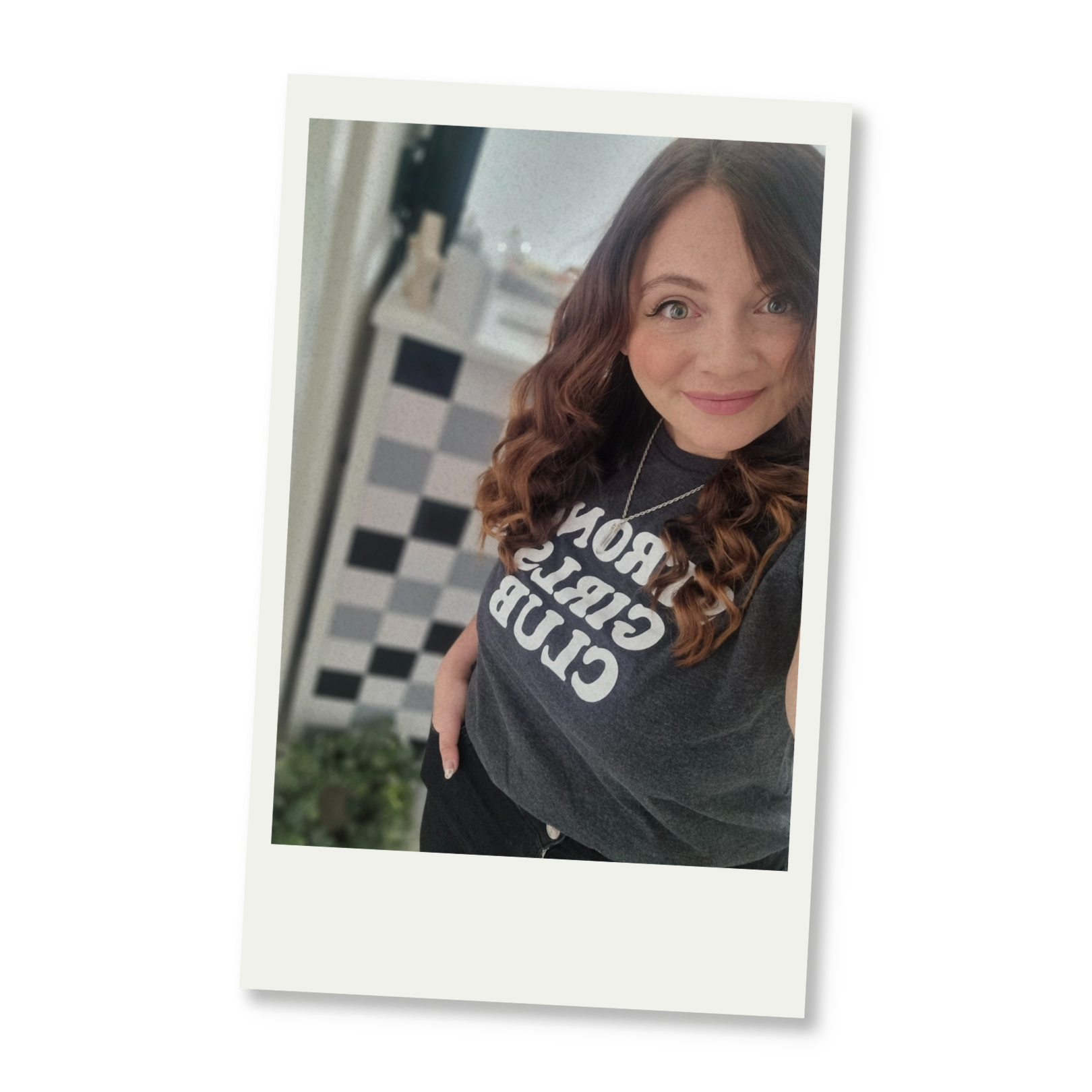 Beth with wavy brown hair and light skin smiling at the camera, wearing a dark T-shirt with white text, standing in front of a checkered wall.