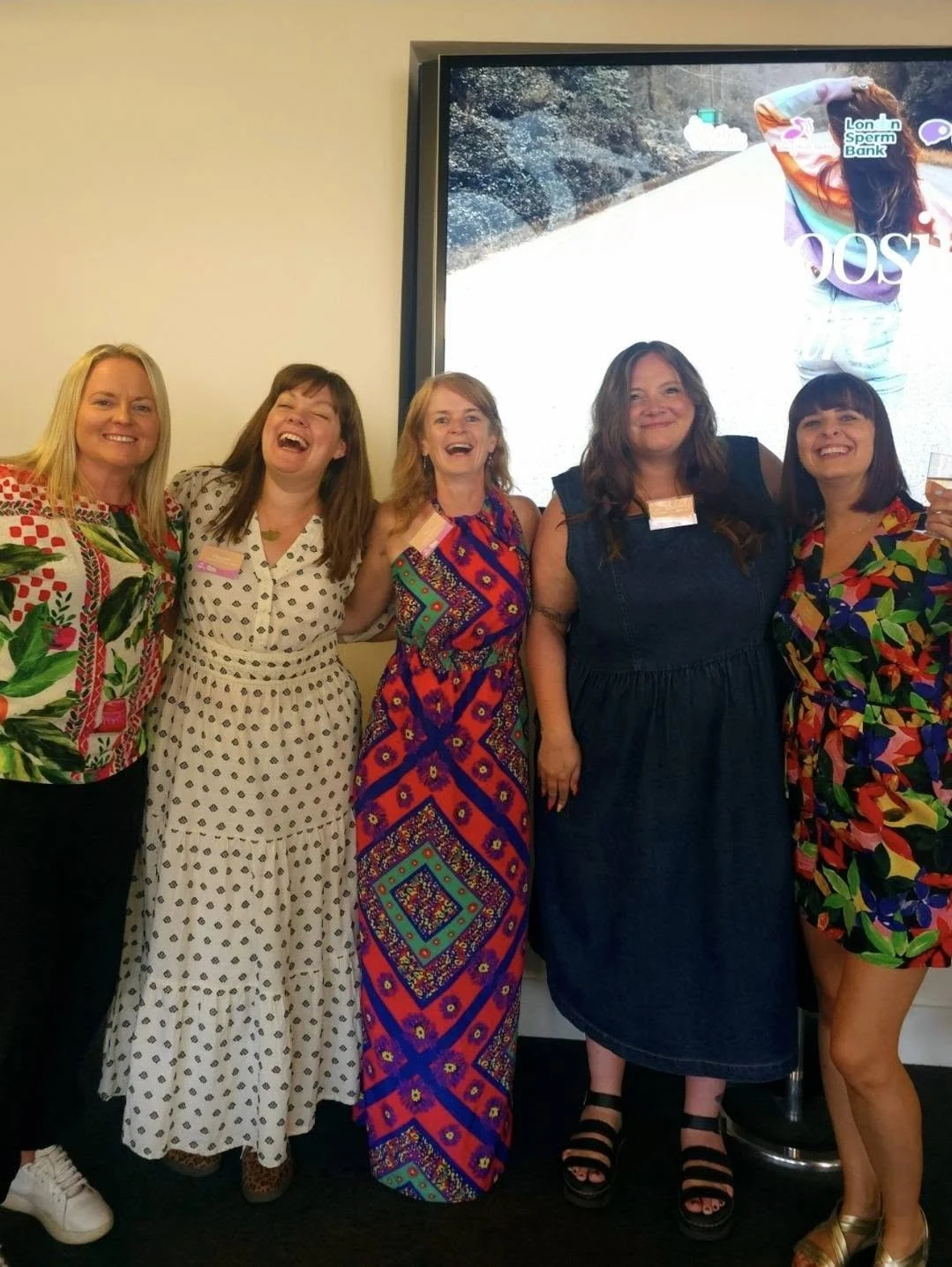 A group of five women standing together, smiling and laughing, at an indoor event. They are dressed in colorful and patterned dresses, with a large screen or monitor behind them displaying a woman sitting outdoors.