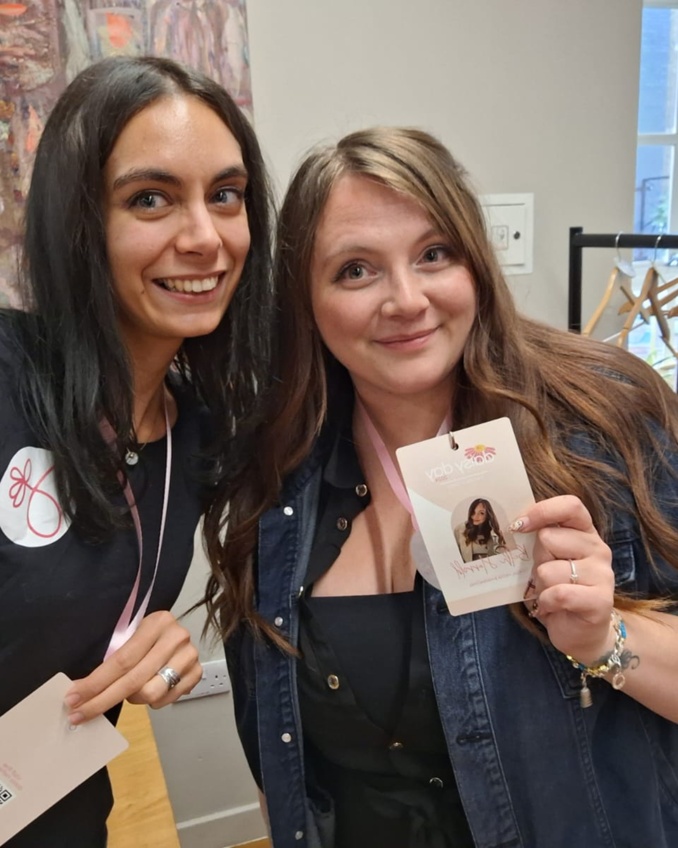 Two women smiling and holding photos or cards, standing indoors with artwork and a window in the background.