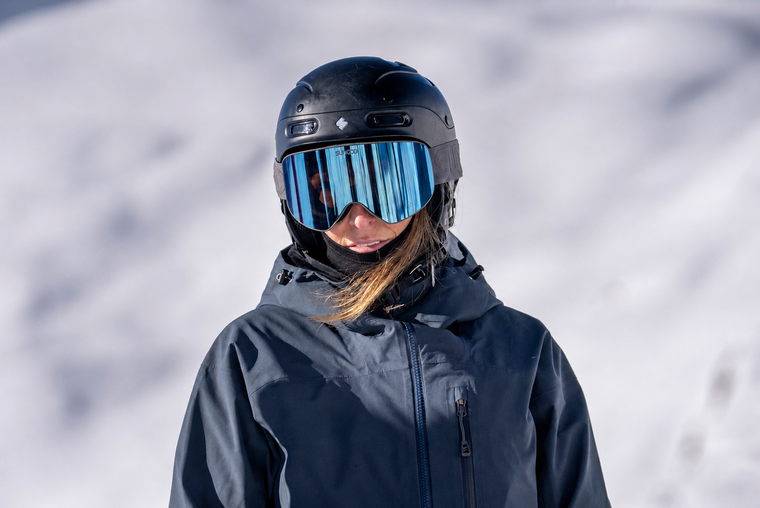 A woman wearing a black ski helmet, a reflective ski mask, and a dark winter jacket standing on snow.