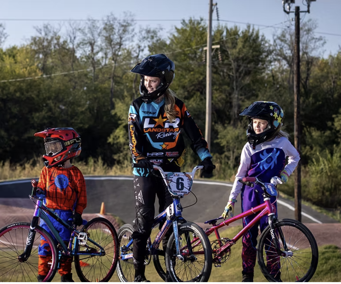 Three children with bicycles and helmets, standing outdoors on a race track with trees and power lines in the background.