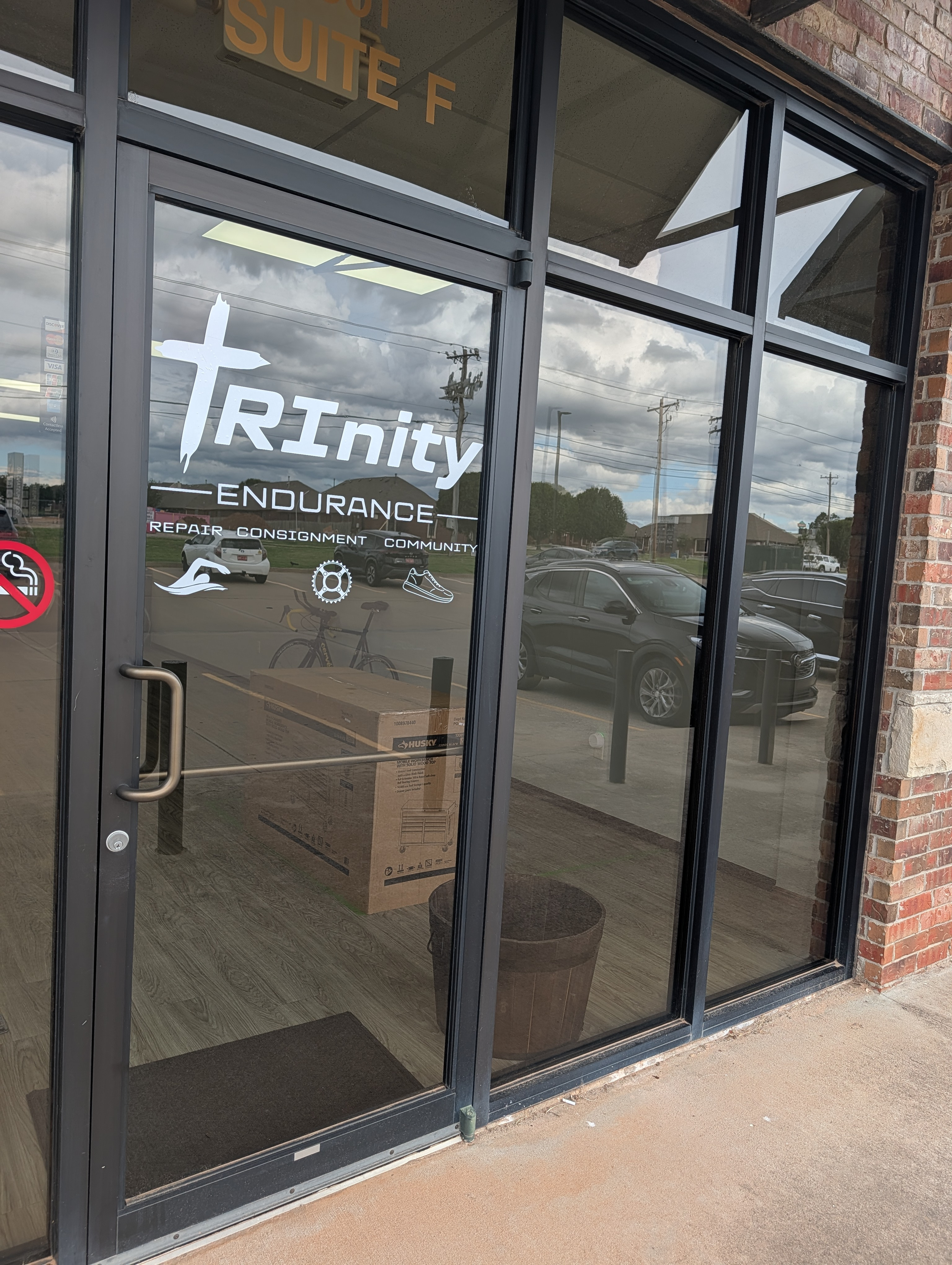 Glass entrance door with the sign 'Trinity Endurance' and icons representing swimming, biking, running, and community support, outside view with parking lot and cloudy sky.