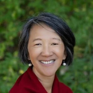 Portrait of a woman with short black hair, smiling, wearing a red top and pearl earrings, outdoors with green foliage in the background.