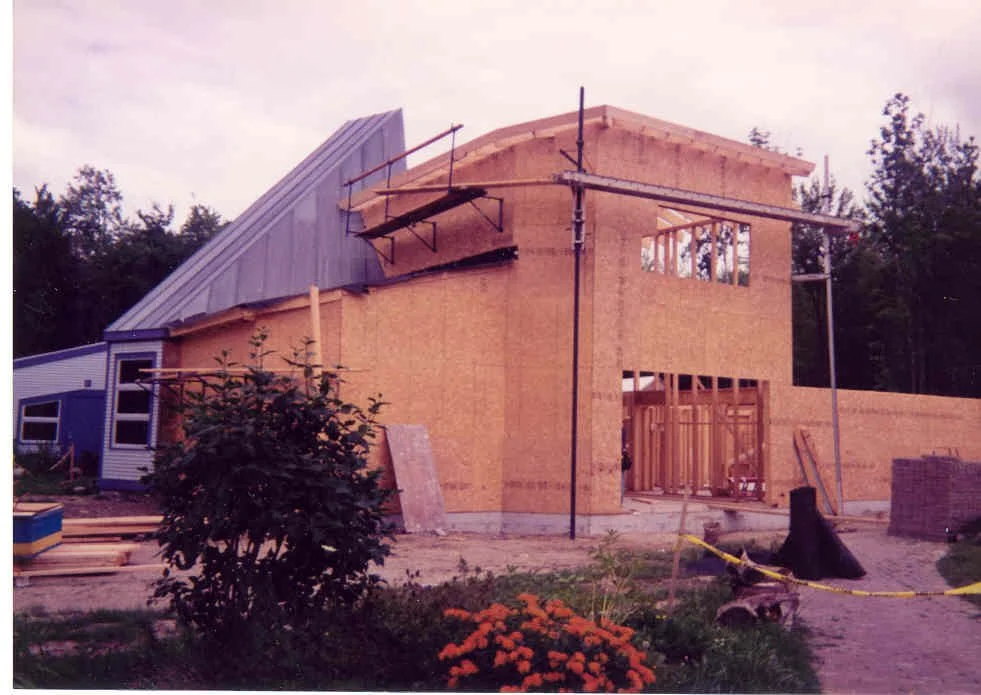 Construction site of a house with wooden framing and partial exterior walls, scaffolding, and building materials around, surrounded by trees and a garden with flowers.