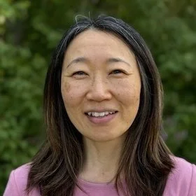 Close-up of a smiling woman with straight, shoulder-length dark hair, wearing a pink top, outdoors with green foliage in the background.