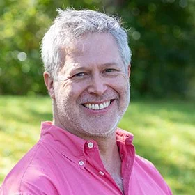 Smiling man with gray hair wearing a pink button-up shirt, outdoors with greenery in the background.
