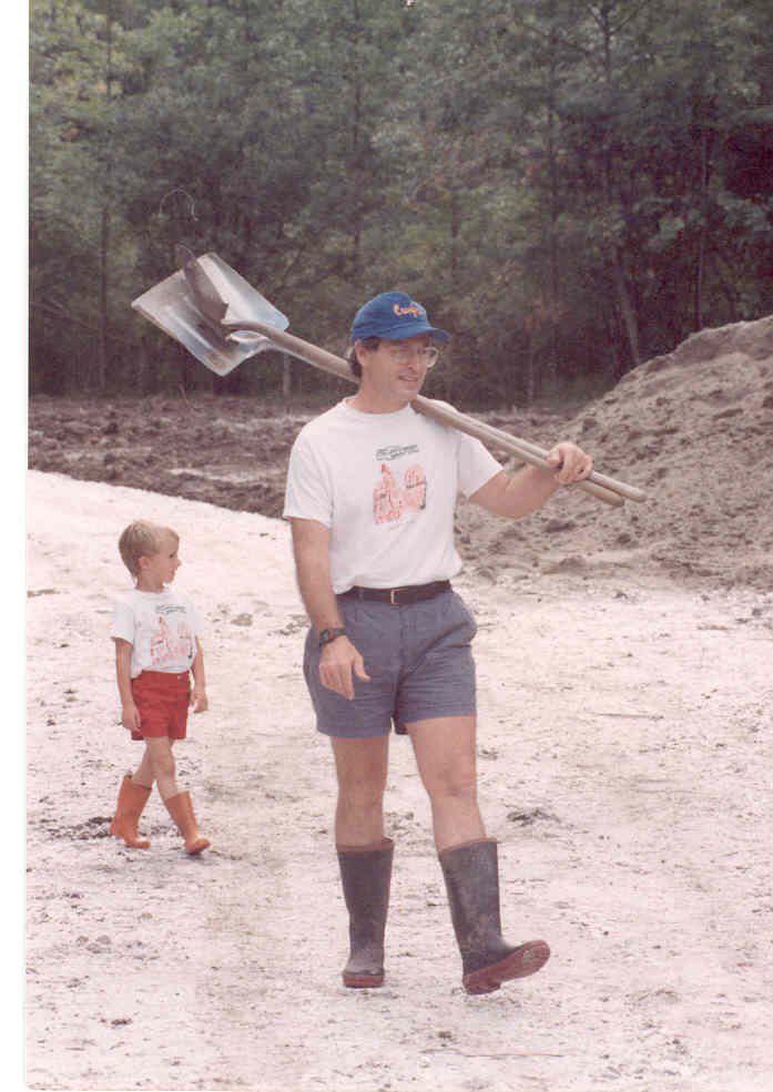 Person carrying a shovel over their shoulder walking on a dirt surface with a child in a forested area in the background.