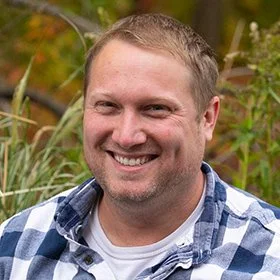 A smiling person with short light brown hair and a beard, wearing a blue and white checkered shirt, outdoors with greenery in the background.