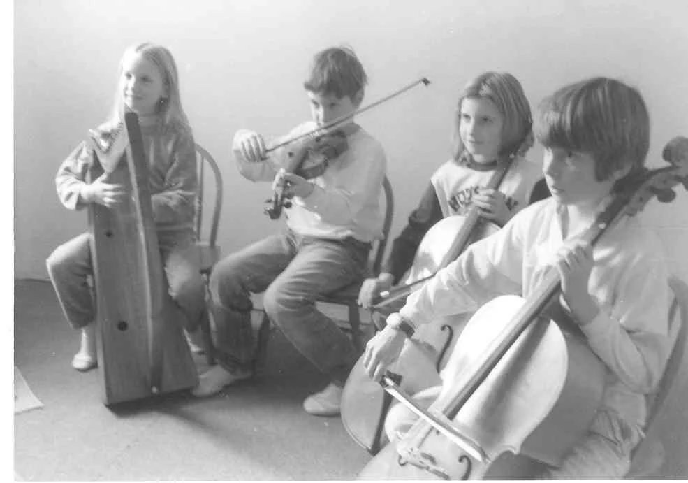 Four children sitting on chairs, playing musical instruments, including a harp, violin, cello, and flute, in a room with plain walls.