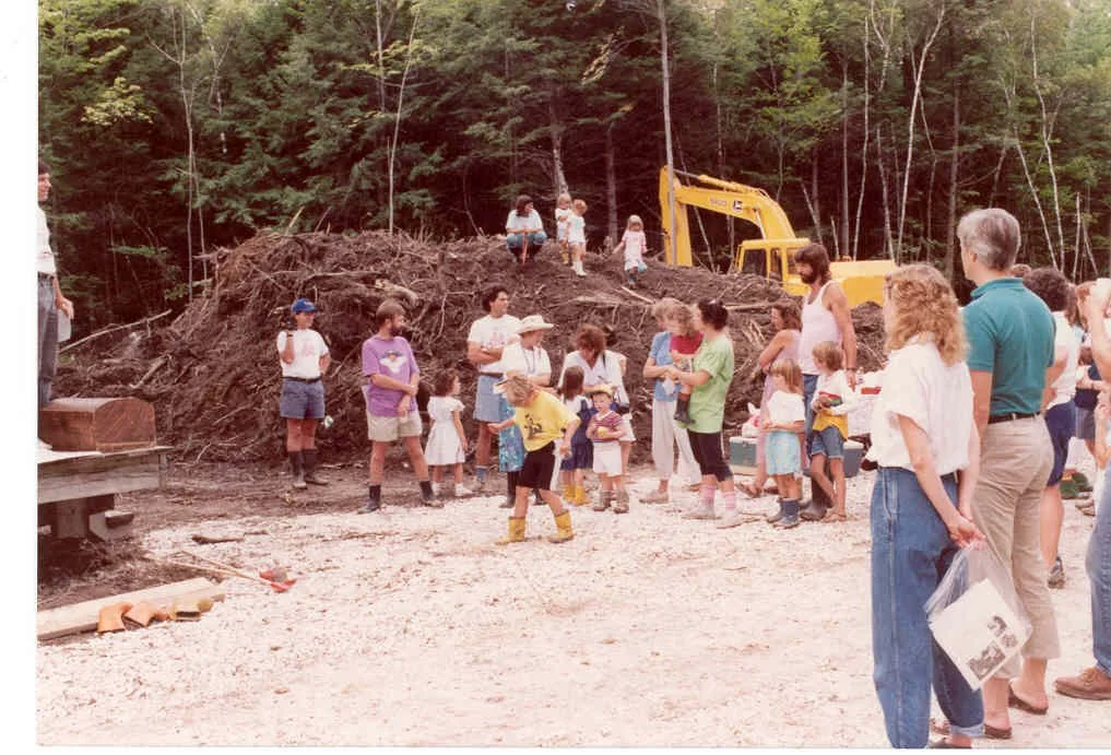 Children and adults gather outdoors near a dirt hill with a yellow excavator, trees in the background, seemingly during a community event or outdoor activity.