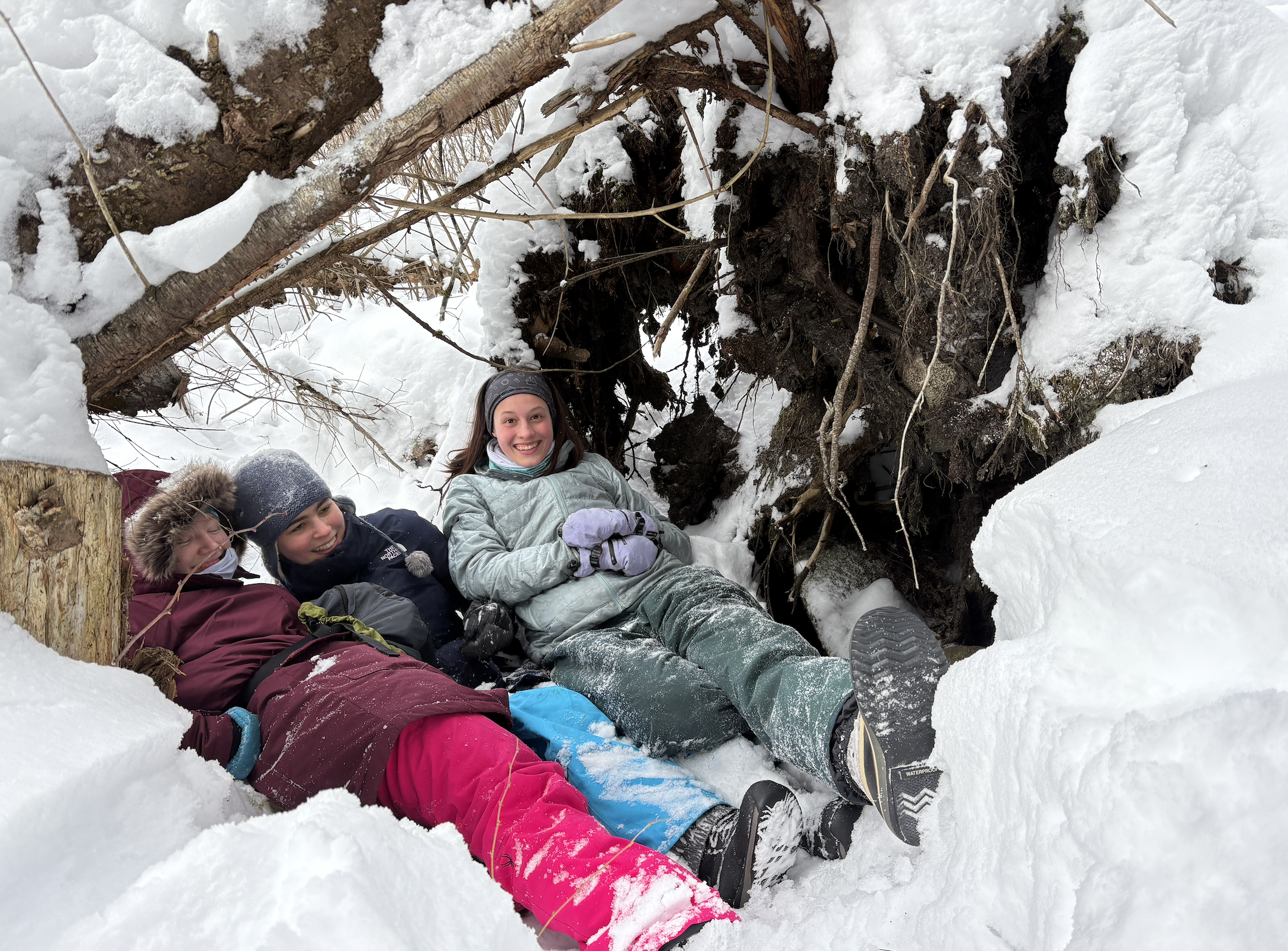 Group of children lying in the snow under an uprooted tree, smiling and wearing winter clothing and boots during a snowy day.