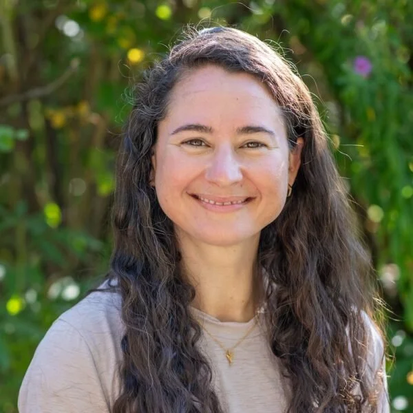 A woman with long, wavy brown hair and a light grey t-shirt smiling outdoors with greenery in the background.