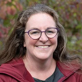 A woman with long wavy gray hair, wearing glasses and a maroon jacket, smiling outdoors with trees and plants in the background.