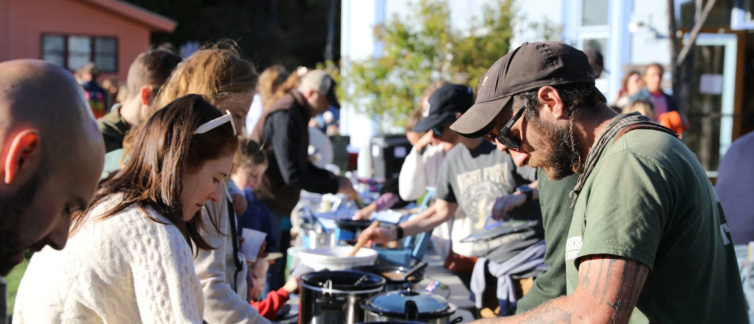 People lined up at a outdoor food stand, serving food from large pots, with a background of trees and buildings.