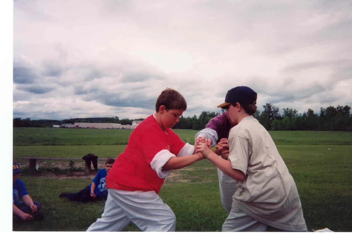 Two boys engaging in a friendly wrestling match outdoors on a grassy field, with a few other children sitting in the background under cloudy skies.
