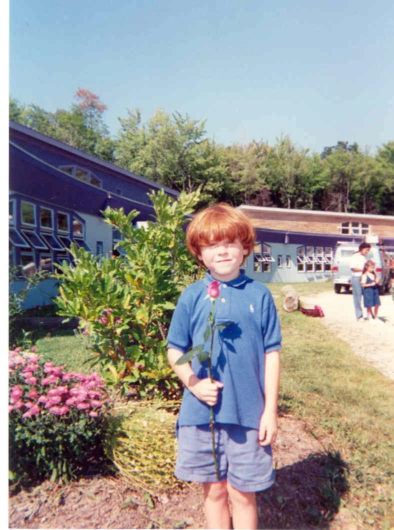 A young boy with red hair wearing a blue polo shirt and shorts holding a pink flower, standing outdoors near a garden with pink flowers and greenery, with buildings and an adult in the background.
