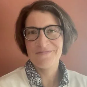 A woman with short brown hair, glasses, and a patterned scarf, smiling in front of a pinkish background.