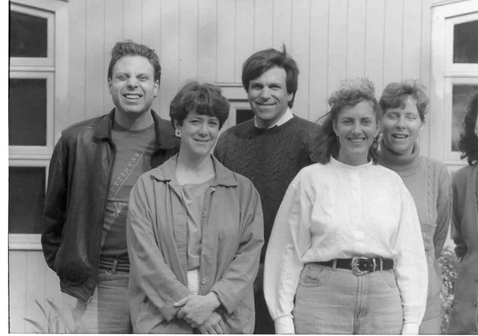 Black and white photo of six people smiling and standing outdoors near a wooden building with windows.