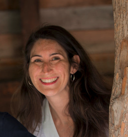 Woman with long dark hair smiling near a tree, background wood beams.