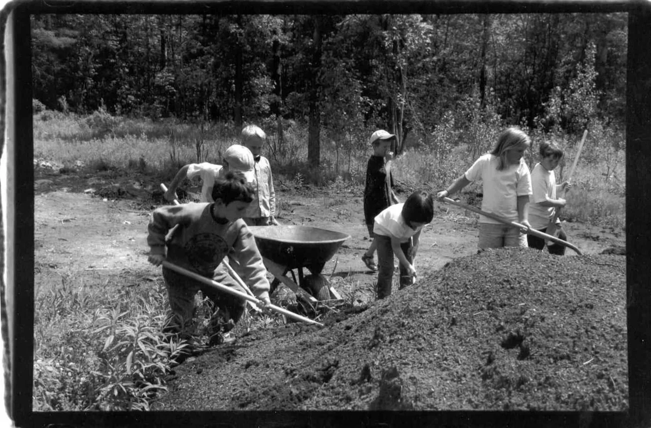 Children playing outside, shoveling dirt onto a large pile, with a wheelbarrow nearby, in a wooded area.