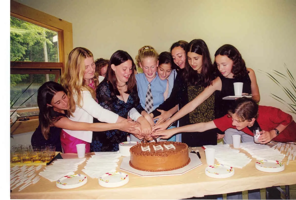 Group of young women and one young man cutting a large birthday cake together at a party.