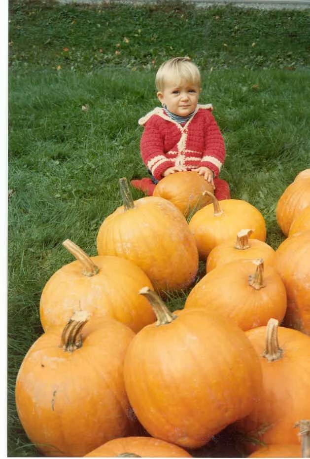 A young child in a red sweater sitting behind a large pile of pumpkins on green grass during the daytime.