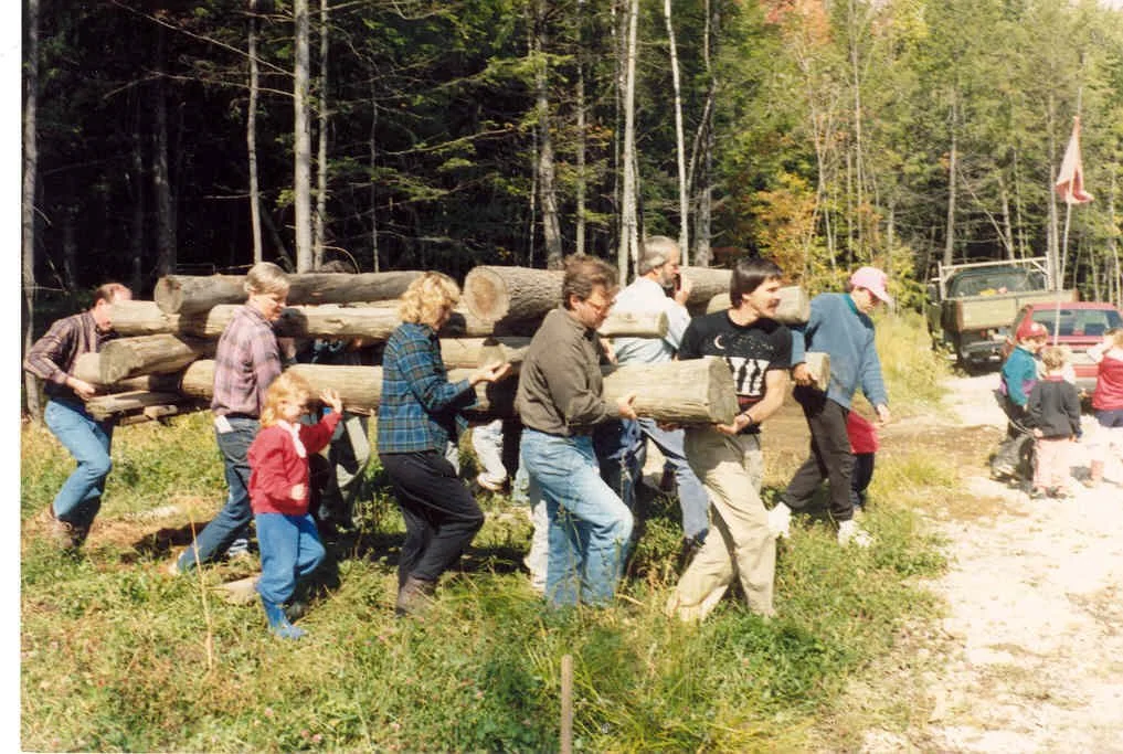 Group of people carrying logs in a forested outdoor area.