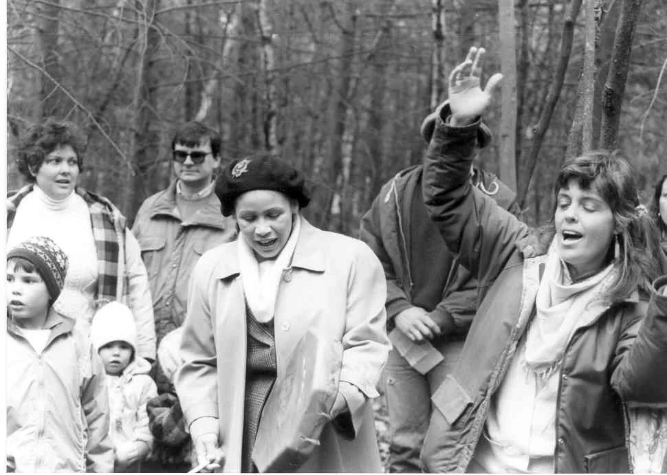 A group of people, including children, in a wooded outdoor setting, with some individuals appearing engaged in a discussion or storytelling, and one woman gesturing with her hand.