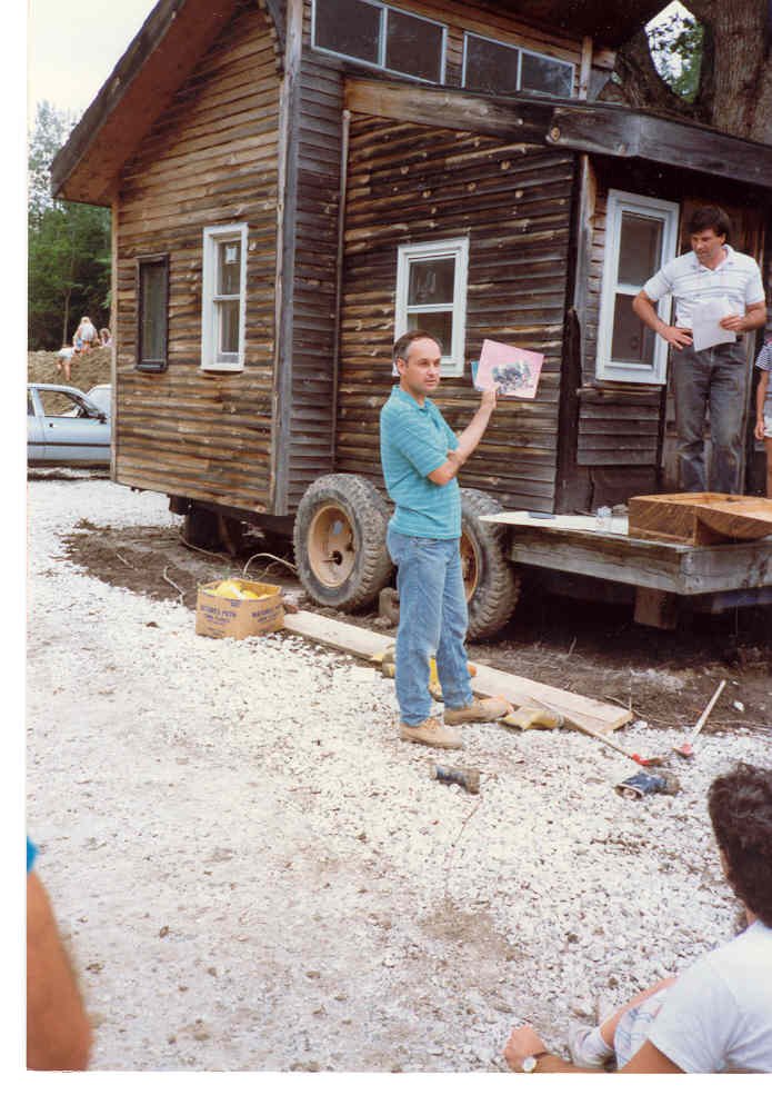Man in blue shirt holding a drawing in front of a tiny house on wheels, with another man standing on the porch reading from a paper, and a woman in the background near a car.