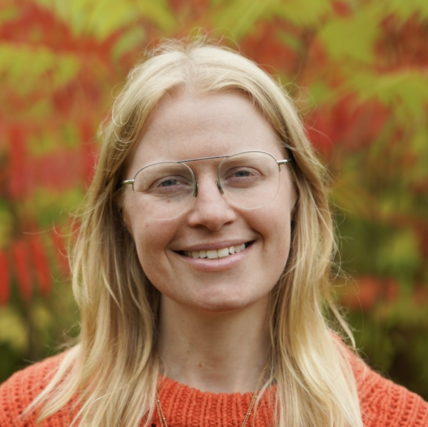 A woman with blonde hair and glasses smiling outdoors with colorful autumn leaves in the background.