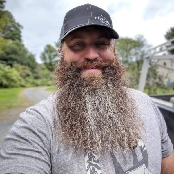 A man outdoors, wearing a black baseball cap with 'Sterling Homes' logo, a gray t-shirt, and smiling with a long, full beard. Trees and a pathway are in the background.