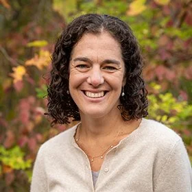 Smiling woman with dark curly hair in a beige sweater outdoors with colorful fall foliage in the background.
