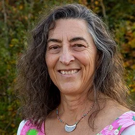 Close-up of a smiling woman with long, curly gray hair outdoors, wearing a pink and purple top and a silver necklace.