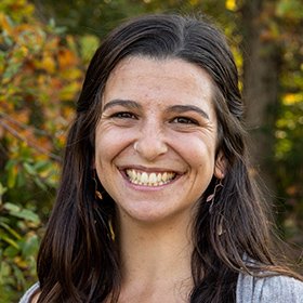 A woman with long, dark hair smiling outdoors with autumn foliage in the background.