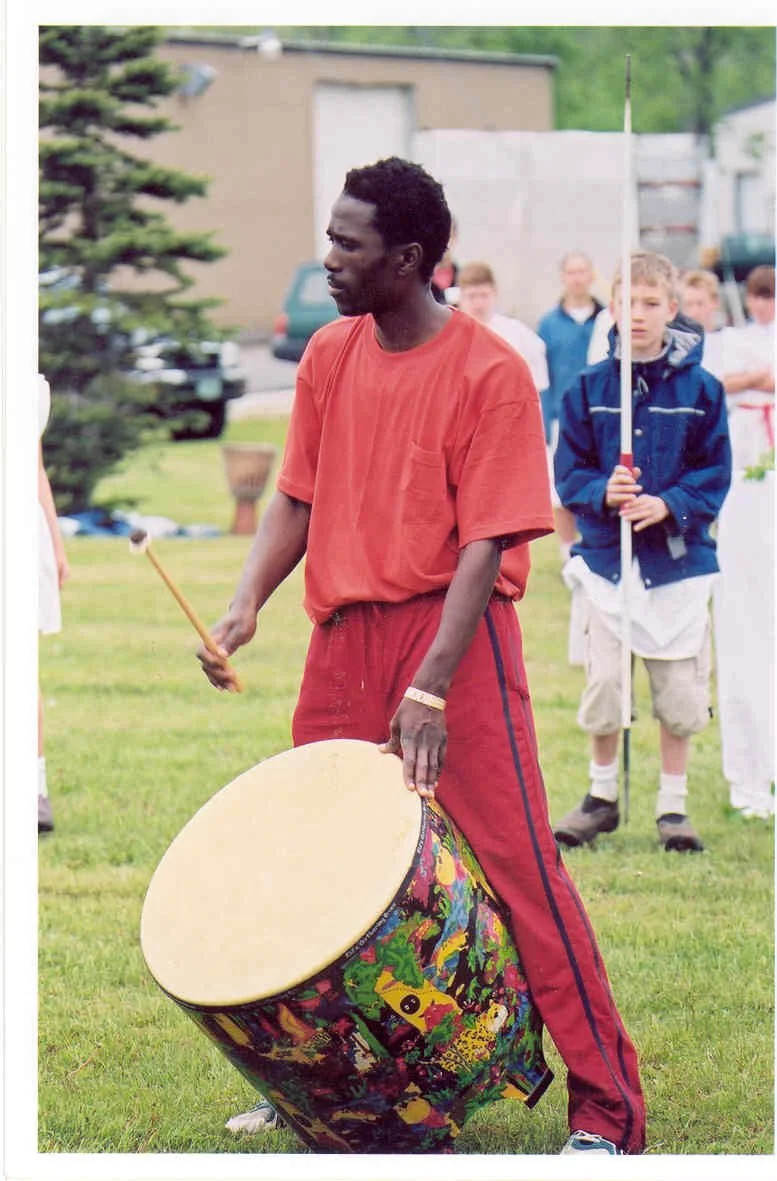 A man playing a large, colorful drum outdoors in a grassy area, with children in the background holding flags and dressed casually.