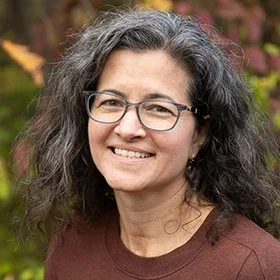Portrait of a woman with gray curly hair and glasses, smiling outdoors with colorful foliage in the background.